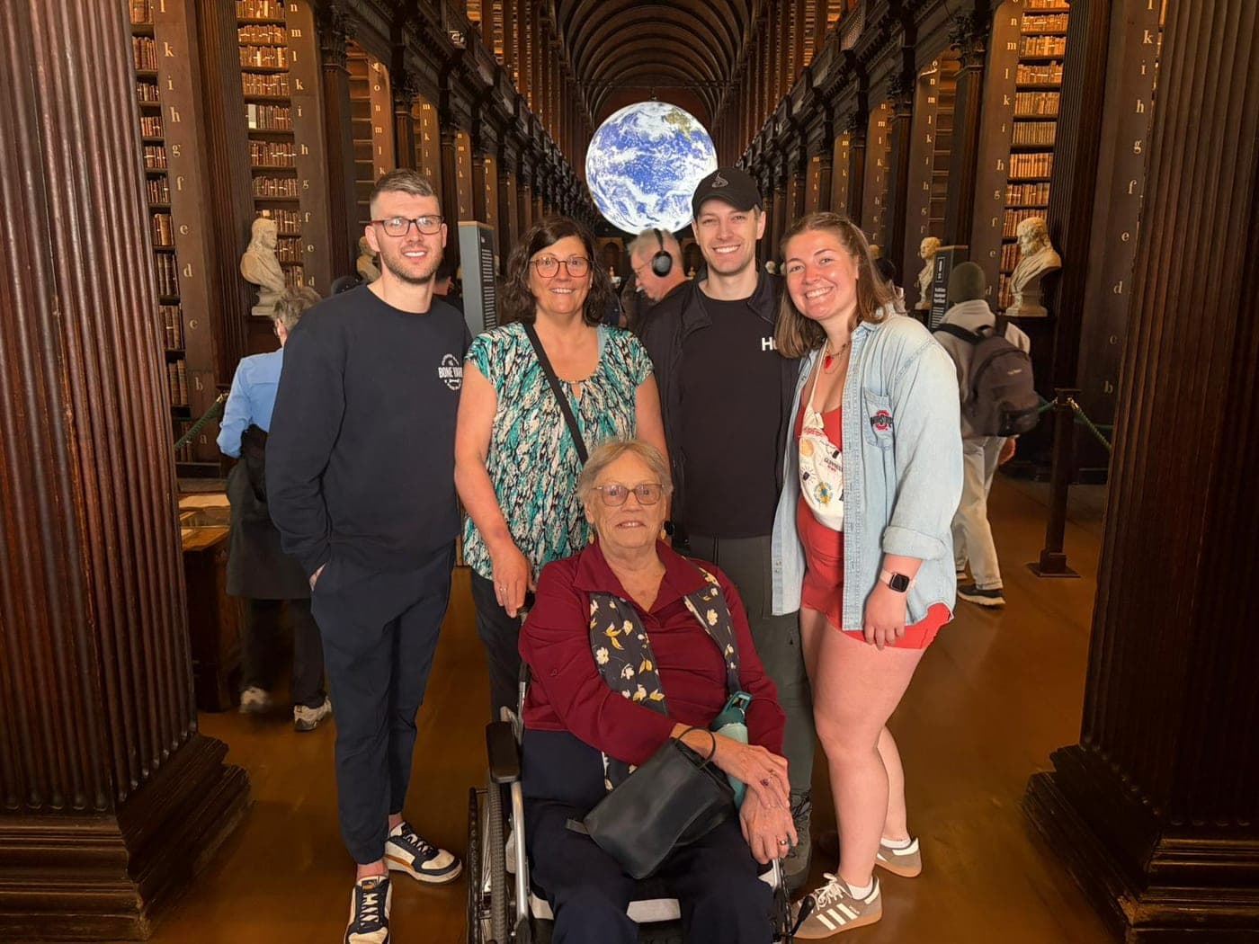 Multi-generational family of five at Trinity College Dublin's Long Room library with grandmother in rented wheelchair, Gaia Earth installation overhead